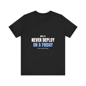 Puede incluir: Una camiseta negra de manga corta con el texto en blanco RULE #1 NEVER DEPLOY, el texto en azul claro ON A FRIDAY y el texto más pequeño en cursiva blanca ask me how I know centrado en el pecho. La camiseta tiene cuello redondo.