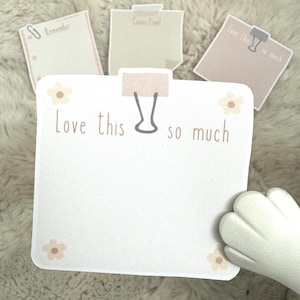 May include: A white note pad with a brown paperclip and the text "Love this so much" with small brown flowers on the corners. The note pad is on a fuzzy white surface.