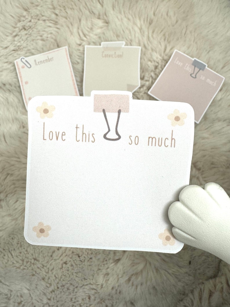 May include: A white note pad with a brown paperclip and the text "Love this so much" with small brown flowers on the corners. The note pad is on a fuzzy white surface.