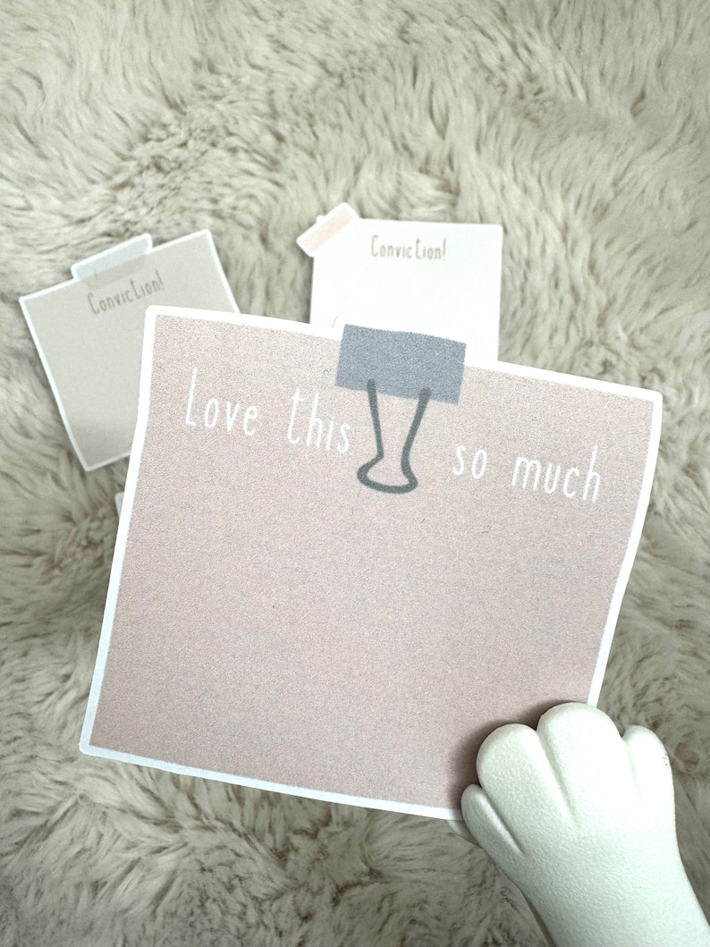 May include: A pink sticky note with the text "Love this so much" written on it. A silver paper clip is attached to the note. The note is on a white fluffy surface.