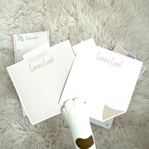 May include: Two white notepads with the text "Conviction!" on them. A white cat paw is holding the notepads. The notepads are on a light brown fuzzy surface.