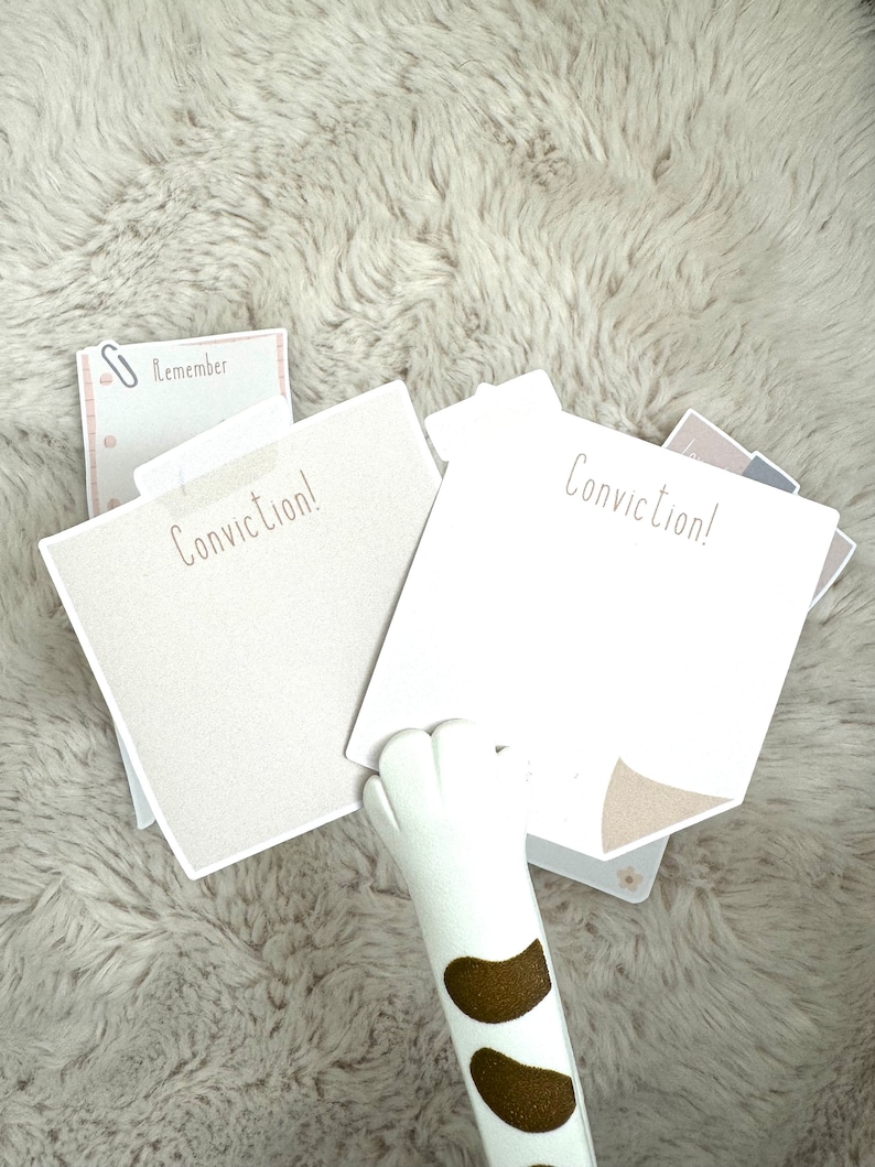May include: Two white notepads with the text "Conviction!" on them. A white cat paw is holding the notepads. The notepads are on a light brown fuzzy surface.