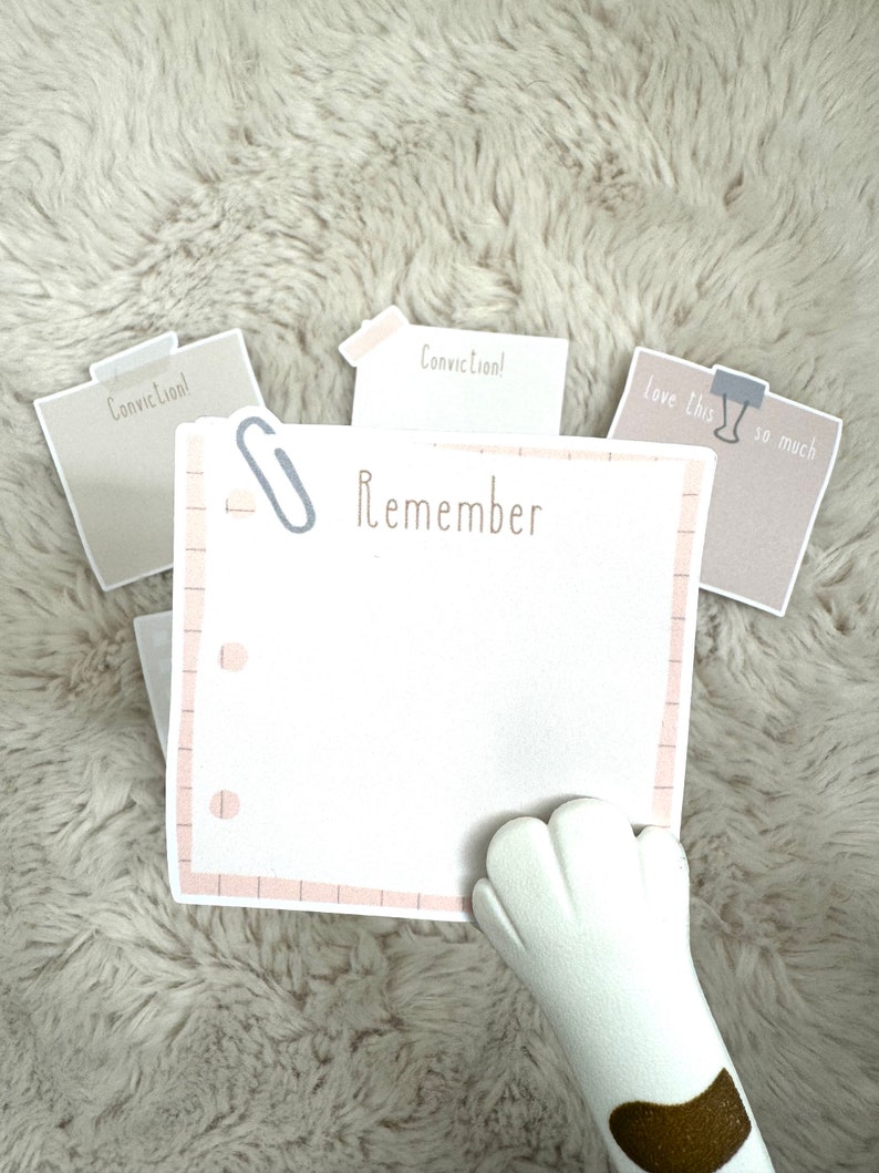 May include: A white notepad with a pink grid pattern and the word "Remember" written in brown text. A silver paperclip is attached to the top of the notepad. Three beige sticky notes with the word "Conviction!" written on them are visible in the background.