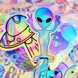 Op de afbeelding: Een set met regenboogkleurige holografische stickers met een alien, een ruimteschip en een cartoonfiguur. De stickers zijn perfect om laptops, waterflessen of dagboeken een vleugje speelsheid te geven.