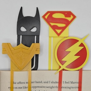 May include: Four superhero-themed bookmarks: a black Batman mask, a yellow Wonder Woman emblem, a red and yellow Superman logo, and a red and yellow Flash emblem. The bookmarks are inserted into a book with visible text.