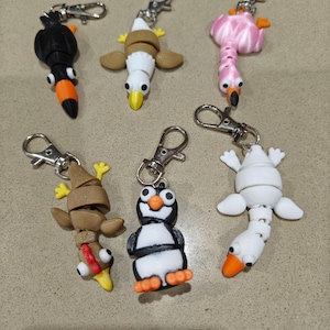 May include: Six bird-shaped keyrings are displayed. The collection includes a black toucan, a brown and white eagle, a pink flamingo, a brown bird with a red beak, a penguin, and a white bird with an orange beak. Each keyring has a silver clasp.