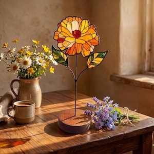May include: A stained glass flower sculpture featuring orange and yellow petals with green leaves, mounted on a circular wooden base. The flower is constructed with dark metal soldering lines between the glass segments, creating a vibrant floral display.