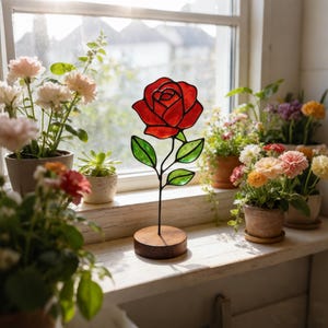 May include: A stained glass rose sculpture featuring a vibrant red flower head and green leaves, held by a thin black metal stem attached to a circular wooden base. The piece is positioned upright, showcasing clear glass segments outlined in dark solder.