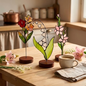 May include: Three stained glass flower sculptures on circular wooden bases. The left piece features red and orange blossoms, the centre piece depicts white lily of the valley, and the right piece shows pink flowers, all with green glass leaves.