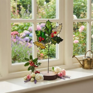 K&ouml;nnte beinhalten: Ein Stechpalmen-Zierzweig aus Buntglas mit gr&uuml;nen und gelben Bl&auml;ttern sowie roten Beeren, montiert auf einem runden Holzsockel. Das Objekt ist umgeben von frischen wei&szlig;en und rosa Rosen, Lavendelzweigen und einem kleinen echten Stechpalmenzweig.