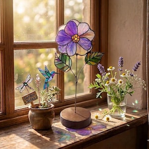 May include: A stained glass purple flower on a wooden base, a small ceramic pot containing a stained glass dragonfly, a hummingbird, a crystal, and a wooden sign labelled Garden Notes, and a glass vase filled with lavender and chamomile flowers.