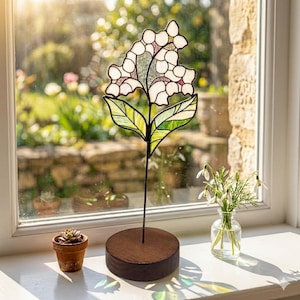 May include: A stained glass flower ornament featuring white and pale pink petals with green leaves, supported by a thin metal stem on a circular dark wood base. The glass pieces are joined by dark solder lines, creating a floral silhouette.