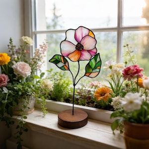 May include: A stained glass flower ornament featuring translucent white, pink, and orange petals with a dark centre, supported by a thin black metal stem and two green leaves, all mounted on a circular wooden base for tabletop display.