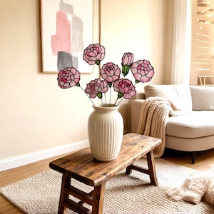 May include: A collection of seven stained glass carnation flowers in shades of pink and white with dark outlines, each attached to a thin metal stem. The flowers are arranged in a vertical, ribbed, off-white ceramic vase with a narrow neck.