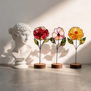 May include: A white classical bust sculpture positioned next to three stained glass flower ornaments on wooden bases. The flowers feature red, pink, and yellow petals with green leaves, each supported by a thin black metal stem on a round base.