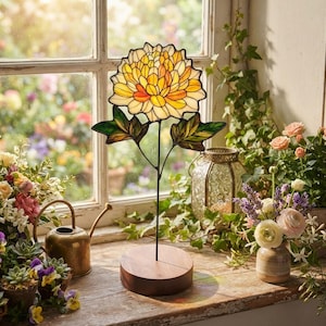 K&ouml;nnte beinhalten: Eine Buntglas-Blumenskulptur mit einer gro&szlig;en gelben und orangefarbenen Chrysanthemenbl&uuml;te und gr&uuml;nen Bl&auml;ttern, montiert auf einem runden Holzsockel. Die Glasbl&uuml;tenbl&auml;tter sind mit dunklem Metall umrandet und bilden ein detailliertes Blumenmuster.