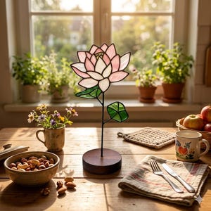 May include: A stained glass lotus flower sculpture featuring light pink and white petals with green leaves, mounted on a thin black metal stem and a circular dark wood base. The flower is constructed with dark metal soldering lines.