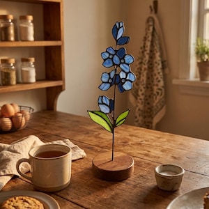 May include: A stained glass flower sculpture featuring blue petals and green leaves, mounted on a circular wooden base. The piece stands upright, showcasing a vertical arrangement of multiple blue blossoms connected by a thin metal stem.