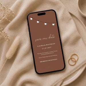 Könnte beinhalten: Ein Smartphone zeigt eine digitale Save-the-Date-Ankündigung auf braunem Hintergrund mit einer Lichterkette aus weißen Lampen. Der Text lautet: save our date, LAURA & MATHIAS, 17.07.2027, sowie weitere Informationen zur Hochzeitszeremonie.