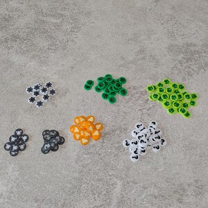 Può includere: Assortimento di pedine da gioco colorate con diversi simboli. Le pedine sono bianche con simboli neri e includono un punto interrogativo, una stella, un cuore, una zucca e una foglia.