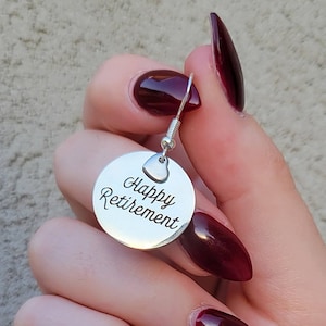 May include: A silver-toned circular metal earring charm featuring the engraved black text Happy Retirement in a cursive script font. The charm is attached to a small silver-toned heart-shaped connector and a standard metal earring hook wire.