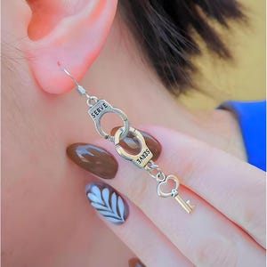 May include: A silver-toned dangle earring featuring two miniature handcuffs linked together, each engraved with the word SERVE, followed by a small chain connected to a heart-shaped key charm at the bottom, suspended from a standard hook wire.