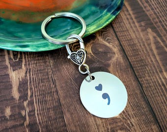 May include: A silver-toned metal keychain featuring a circular disc pendant engraved with a dark gray semicolon and a small heart symbol above it. The disc is attached to a heart-shaped lobster clasp and a standard round split ring for keys.