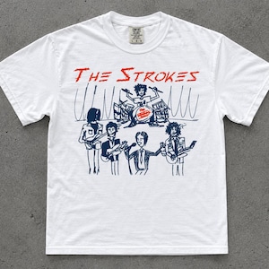 May include: A white short-sleeve t-shirt featuring a dark blue hand-drawn style illustration of a band performing on stage. The text THE STROKES is printed in red above the illustration, and the band name is also on the drum kit in the center.