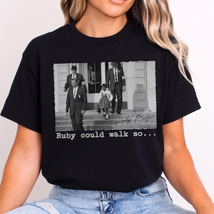 May include: Black t-shirt featuring a black and white image of Ruby Bridges being escorted by U.S. Marshals. The image is framed with the text "Ruby could walk so..." and Ruby Bridges' signature.