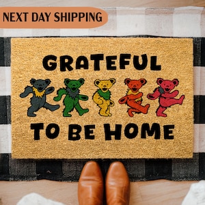 May include: A rectangular doormat with the words "GRATEFUL TO BE HOME" in black. Four colorful dancing bear figures are above the words. The mat is tan with a black and white checkered border.