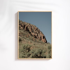 Peut inclure: Une photographie d'un flanc de montagne rocheux avec un ciel bleu en arrière-plan. Le flanc de montagne est recouvert de végétation verte et a une teinte brun rougeâtre.