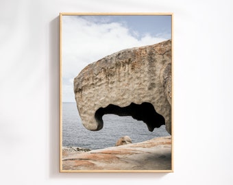 May include: A large, grey rock formation with a natural archway, overlooking a blue ocean. The rock is textured and weathered, with a rough surface. The ocean is calm and the sky is partly cloudy.