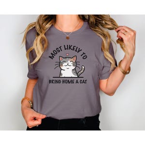 以下が含まれることがあります： 猫の漫画と「Most Likely to Bring Home a Cat」というテキストが書かれたグレーのTシャツ。