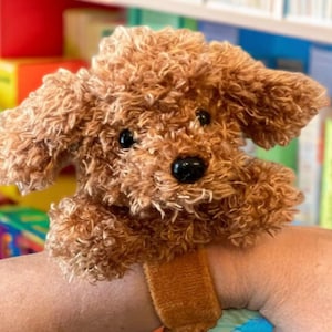 May include: A brown plush toy dog with a furry texture, wearing a brown wristband. The dog is looking at the camera.