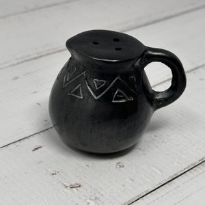 K&ouml;nnte beinhalten: Ein schwarzer Keramik-Krug mit geometrischem Muster und einem kleinen Ausguss. Der Krug hat einen Henkel und drei L&ouml;cher oben.