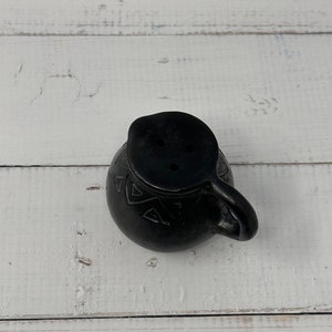 K&ouml;nnte beinhalten: Schwarzer Keramik-Krug mit geometrischem Muster und einem kleinen Ausguss. Der Krug hat einen Henkel und steht auf einer wei&szlig;en Holzoberfl&auml;che.