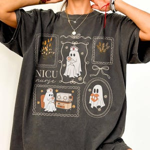 May include: A charcoal gray short-sleeve t-shirt featuring a graphic print with the text NICU nurse. The design includes multiple framed illustrations of ghosts dressed as nurses, a baby, medical equipment, and floral accents in white and tan.