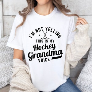 May include: White t-shirt with the text "I'M NOT YELLING THIS IS MY Hockey Grandma VOICE" in black. The design includes crossed hockey sticks and stars. The shirt is a casual wear item.