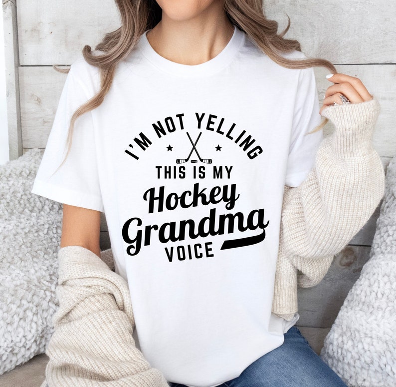 May include: White t-shirt with the text "I'M NOT YELLING THIS IS MY Hockey Grandma VOICE" in black. The design includes crossed hockey sticks and stars. The shirt is a casual wear item.
