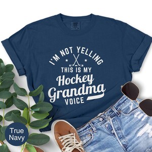 May include: A navy blue t-shirt with the text "I'M NOT YELLING THIS IS MY Hockey Grandma VOICE" in white. The design includes crossed hockey sticks and stars. The shirt is paired with denim shorts, sunglasses, and brown shoes.
