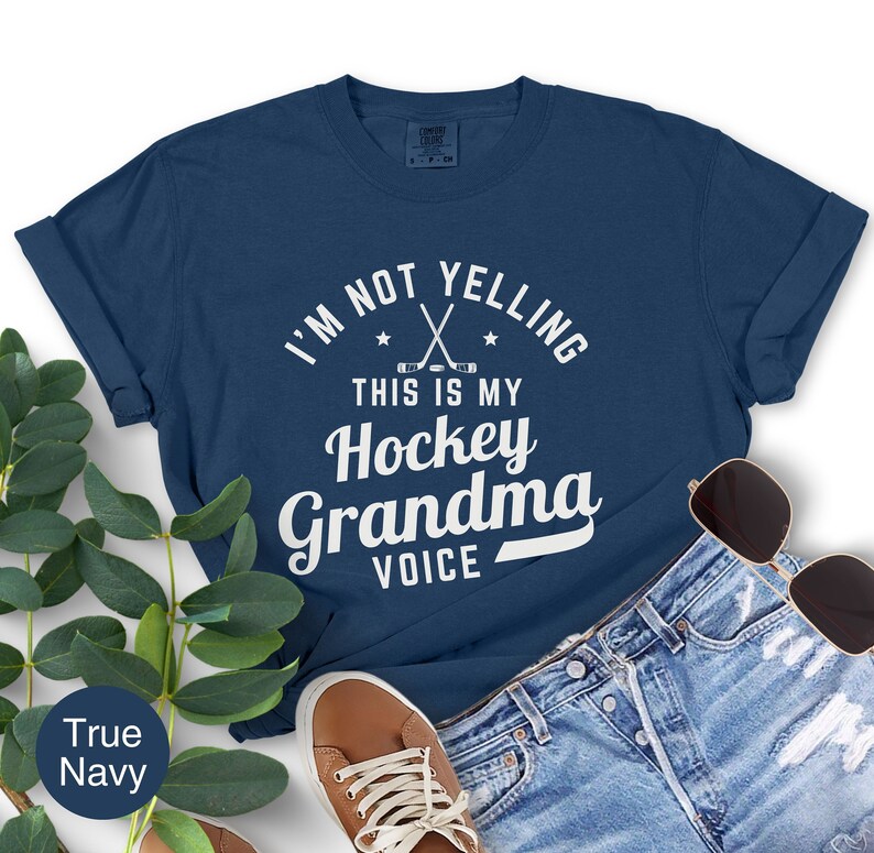 May include: A navy blue t-shirt with the text "I'M NOT YELLING THIS IS MY Hockey Grandma VOICE" in white. The design includes crossed hockey sticks and stars. The shirt is paired with denim shorts, sunglasses, and brown shoes.