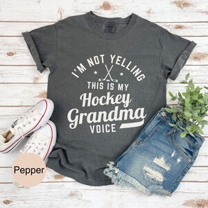 May include: Dark grey t-shirt with the text "I'M NOT YELLING THIS IS MY Hockey Grandma VOICE" in white. The shirt is paired with denim shorts and white sneakers. A sprig of green foliage is visible.