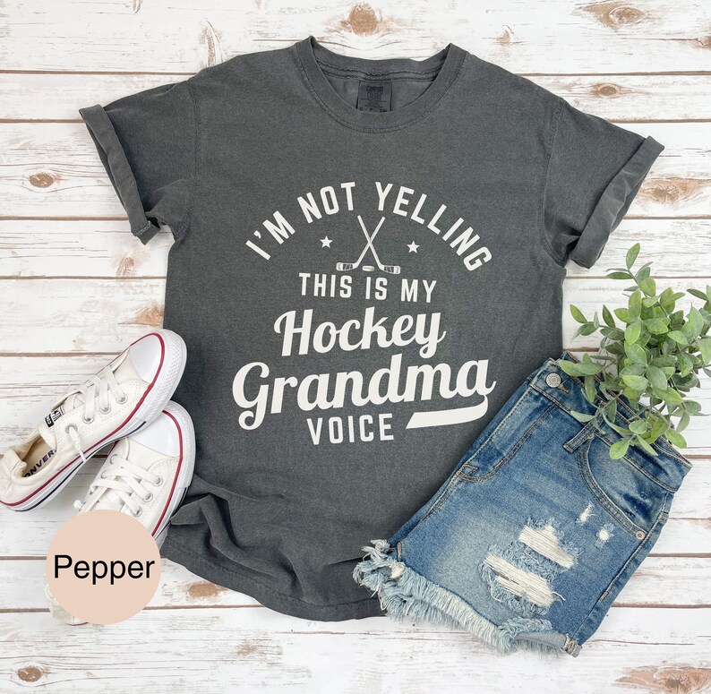May include: Dark grey t-shirt with the text "I'M NOT YELLING THIS IS MY Hockey Grandma VOICE" in white. The shirt is paired with denim shorts and white sneakers. A sprig of green foliage is visible.