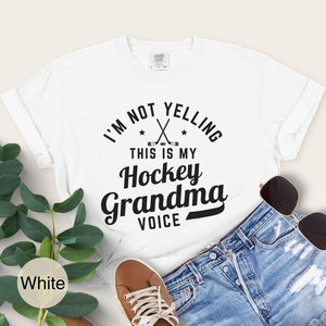 May include: White t-shirt with the text "I'M NOT YELLING THIS IS MY Hockey Grandma VOICE" in black. The shirt features crossed hockey sticks and stars. The shirt is paired with denim shorts, brown shoes, and sunglasses.