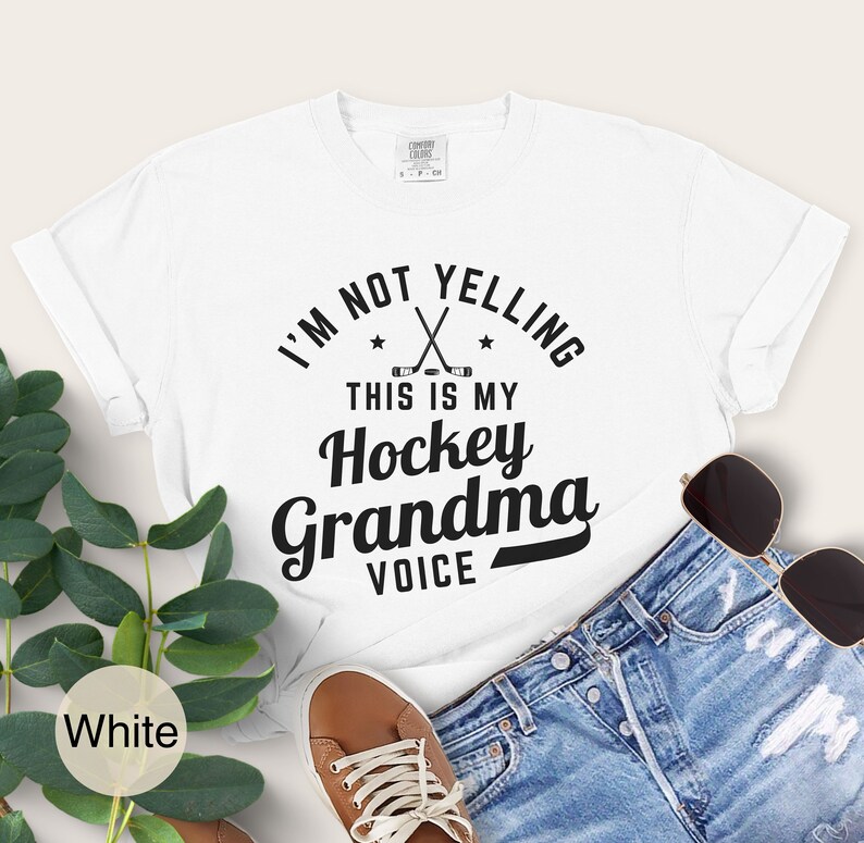 May include: White t-shirt with the text "I'M NOT YELLING THIS IS MY Hockey Grandma VOICE" in black. The shirt features crossed hockey sticks and stars. The shirt is paired with denim shorts, brown shoes, and sunglasses.