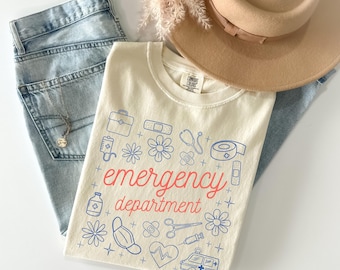 May include: Ivory-colored t-shirt with the words "emergency department" in red, surrounded by blue medical-themed illustrations. The shirt is paired with light blue jeans and a tan hat.