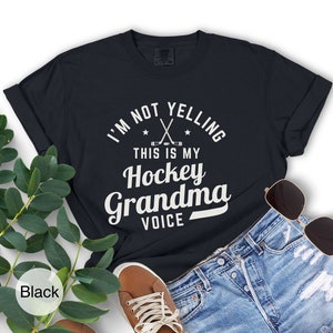 May include: Black t-shirt with the text "I'M NOT YELLING THIS IS MY Hockey Grandma VOICE" in white. The design includes crossed hockey sticks and stars. The shirt is paired with denim shorts, sunglasses, and a brown shoe.