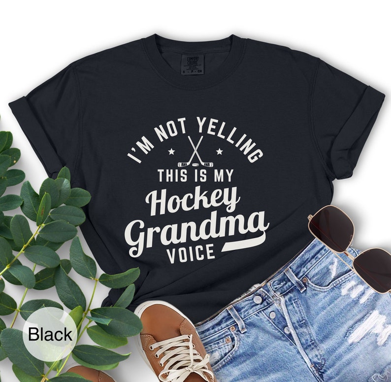 May include: Black t-shirt with the text "I'M NOT YELLING THIS IS MY Hockey Grandma VOICE" in white. The design includes crossed hockey sticks and stars. The shirt is paired with denim shorts, sunglasses, and a brown shoe.