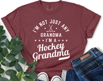 May include: A maroon t-shirt with white text that reads "I'm not just any grandma I'm a hockey grandma". The text is surrounded by stars and a graphic of two hockey sticks crossed.