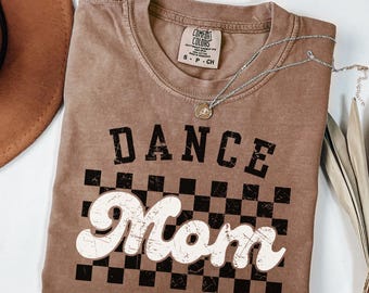 Op de afbeelding: Espresso-kleurig t-shirt met de woorden "DANCE Mom" in een verweerde lettertype over een zwart-wit geblokt patroon. Een zilveren ketting met een hanger is over het shirt gedrapeerd.